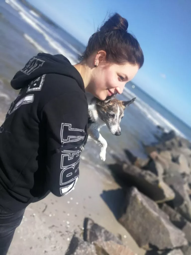 Katja lächelnd mit ihrem Hund am Sandstrand des Meeres